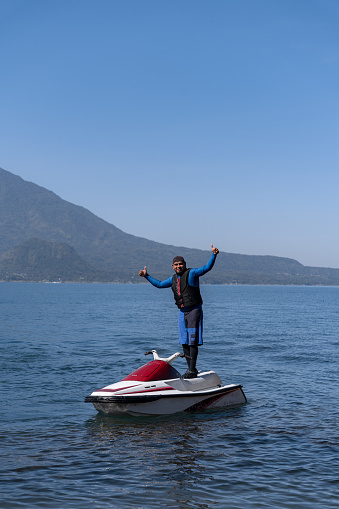 A young Hispanic man demonstrates stability and control while standing on a jet-ski, set against the scenic landscape of Atitlan Lake in Guatemala
