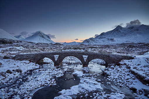 Sligachan old Bridge, Isle Of Skye, Scotland. Sligachan bridge during sunset, the water flows quickly under the bridge. In the background you can see the mountains on Skye that are covered in snow.