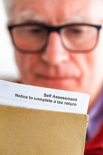 Senior self-employed man opening a tax letter from HMRC in a brown envelope. Focus on the letter with the man defocused in the background.
