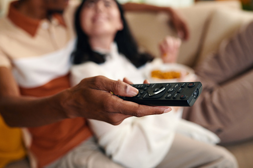 Hand of young African American man with remote control choosing channel while sitting next to his wife in front of TV set at home