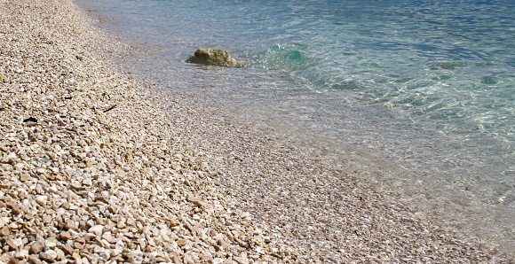 Small Pebbles On The Seashore. Small Pebbles On Beach. Clear Sea Water. Blue Mediterranean Sea.
