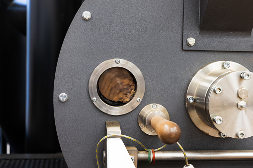 Industrial coffee roaster close-up photo, roasting coffee seeds are rotating inside behind small round technological window