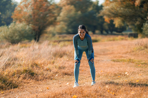 A smiling athletic woman standing with hands on her knees after her run in nature.