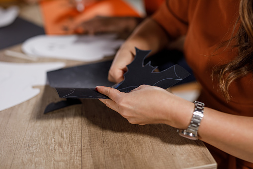 Close up shot of woman and her two teenage sons cutting out bats and decorations for Halloween out of paper when doing crafts together.