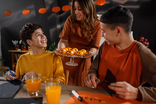 Caring mother serving tangerines, with Jack o' Lantern faces drawn on them, to her two teenage boys while they are doing Halloween crafts.