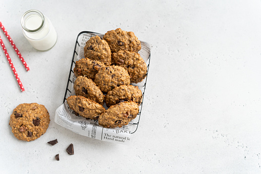 Homemade low-calories banana cookies with oatmeal, dark chocolate drops and walnuts in a metallic basket and a bottle of milk on light background. Copy space. Healthy food. Oatmeat biscuits.