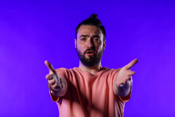 Front view of mid adult hipster standing against bright blue background, gesturing with open arms towards camera as if he is having a discussion with you. Point of view.