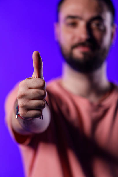Selective focus shot of charming hipster standing against bright blue background, looking at camera, smiling and showing thumbs up.
