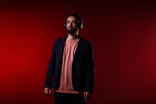 Portrait of charming mid adult man standing against bright red background, listening to music or a podcast via headphones.