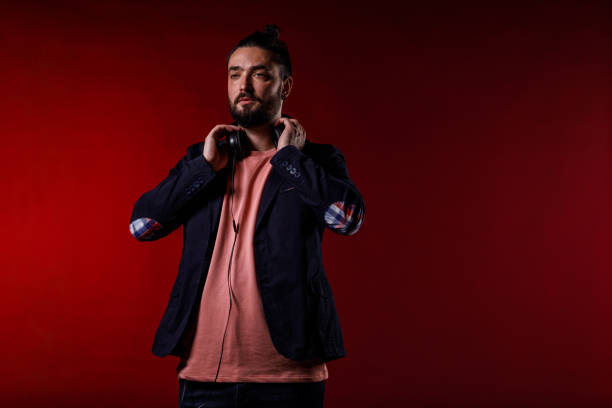 Portrait of charming mid adult man standing against bright red background, putting on headphones to listen to music or a podcast.