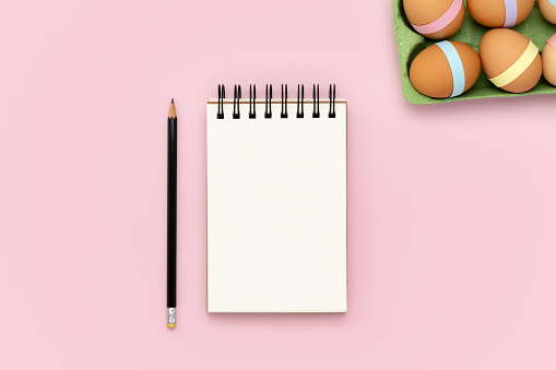 Blank spiral notebook notepad and pencil on pink background with green egg carton and decorated Easter. Flat lay design mockup with empty space. Holiday planning banner