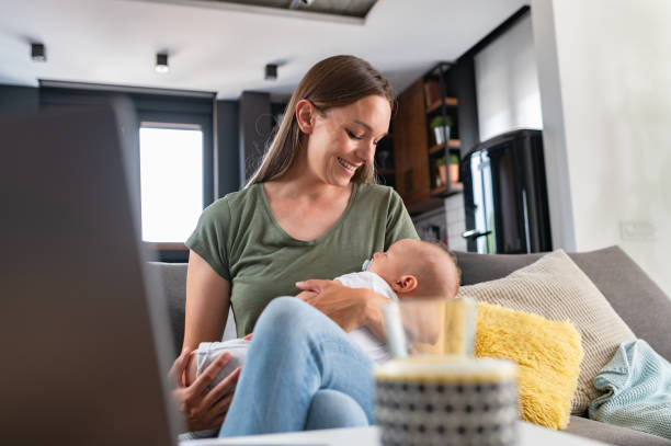 istock Young new mom holding cute baby in her arms at home while working on a computer 1990105910
