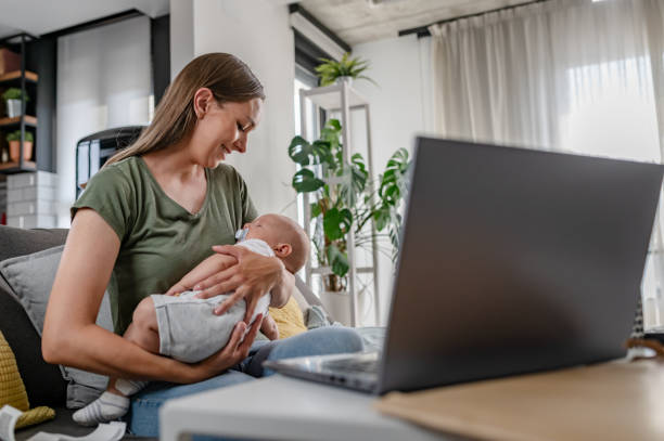 istock Young new mom holding cute baby in her arms at home while working on a computer 1990112130