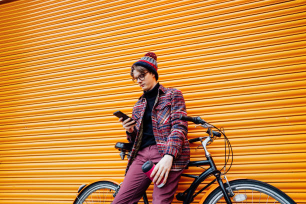 istock Young hipster man using mobile phone while sitting on cruiser bicycle near orange wall. Student using 5g network,rests after riding. Eco friendly transport for carbon footprint. Sustainable lifestyle 1990131778