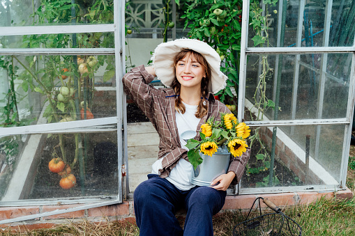 Portrait of smiling young female farmer woman holding watering can with fresh sunflower bouquet, sits near greenhouse with ripe tomatoes. Urban farming lifestyle. Growing organic vegetables in garden