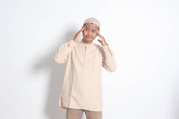 portrait of suffering asian muslim man in white shirt having a migraine, touching his temple. headache disease concept. isolated image on white background - pusing photos et images de collection