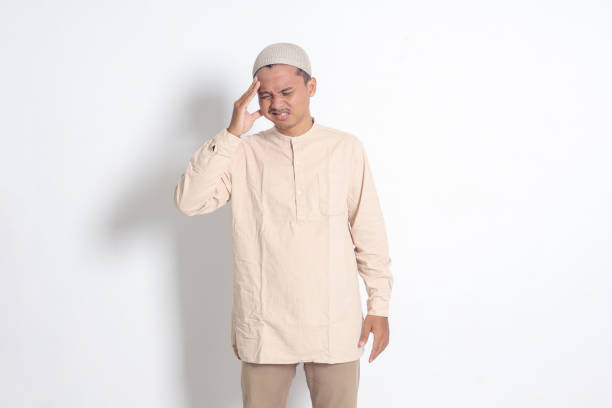 portrait of suffering asian muslim man in white shirt having a migraine, touching his temple. headache disease concept. isolated image on white background - pusing photos et images de collection