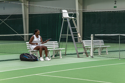 Front view female tennis player sitting on bench while holding tennis racquet in tennis court.