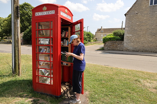 Woman looking through a village community book sharing project in the village of South Cerney in The Cotswolds which breathes life onto an old British red telephone box and keeps an icon of Britain from the past alive.