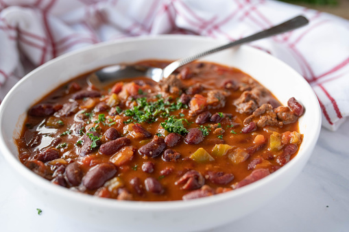 Healthy kitchen with a fresh homemade cooked kidney bean soup or stew with lean ground beef, tomatoes, peppers, chili, onions, garlic and herbs. Served ready to eat on a soup plate with spoon isolated on light background.