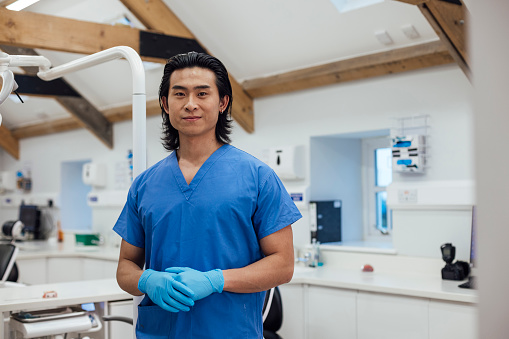 A medium close up of a young male orthodontist smiling and looking into the camera in the practice that he works at in the North East of England. He is dressed in surgical scrubs and gloves with his hands held together