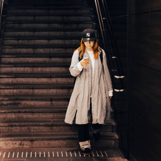 Tokyo, Japan, April 30st, 2023  - Woman wearing a baseball cap and raincoat, holding a mobile phone and  reading the screen, whilst entering a subway entrance staircase.