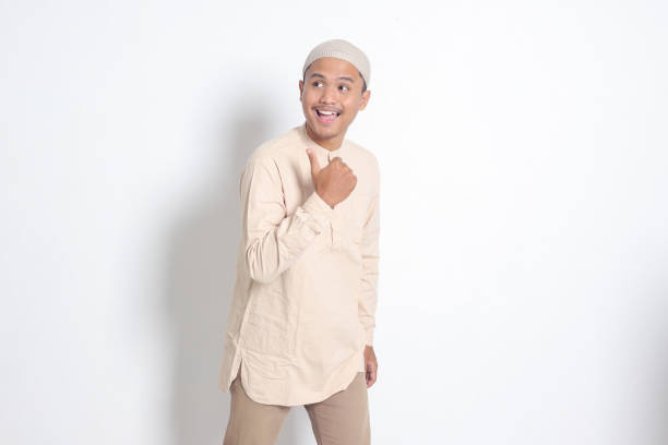 Portrait of shocked Asian muslim man in koko shirt with skullcap showing product and pointing with thumb up to the side. Advertising concept. Isolated image on white background stock photo