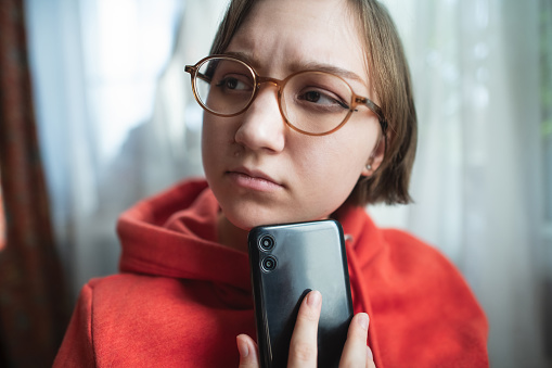 Concerned, sad and worried young woman with smartphone at home reading some bad news or having problems