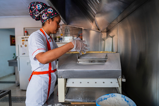 Latina woman employed by a small food business specializing in empanadas, dressed in a uniform, is in the kitchen of the business preparing the implements she uses for the day's production.