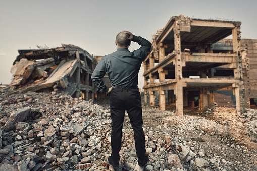 Conceptual image with a businessman holding his head looking at the ruins of a residential area, mixed media