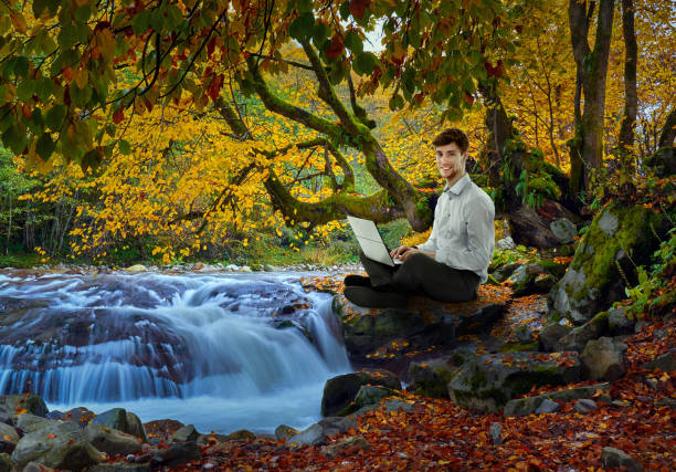 Businessman working on the laptop sitting by the river in a colorful autumnal landscape