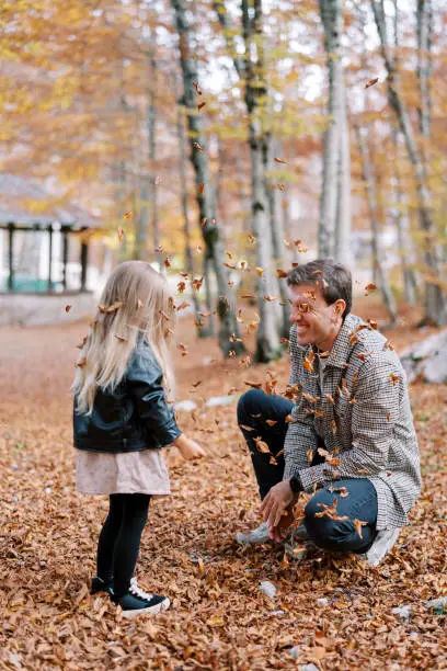 Photo of Little girl stands next to her squatting father under falling dry leaves. Back view