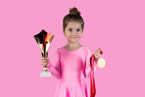 Joyful sports girl celebrating victory isolated on pink background wearing gold medal showing trophy she won. High quality photo