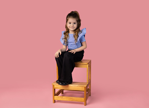 Very stylish and smiling 5-6 year old girl posing in the studio, sitting on a wooden chair, isolated on a pink background. High quality photo