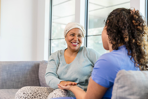 Sitting on the sofa in the living room, the mid adult female cancer patient smiles at the unrecognizable female hospice nurse.