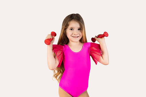 Close-up portrait, child, smiling, funny looking, happy little girl wearing pink gymnastics leotard, lifting dumbbells, isolated white background. Facial expressions, emotions, attitude, determination, dynamism. High quality photo