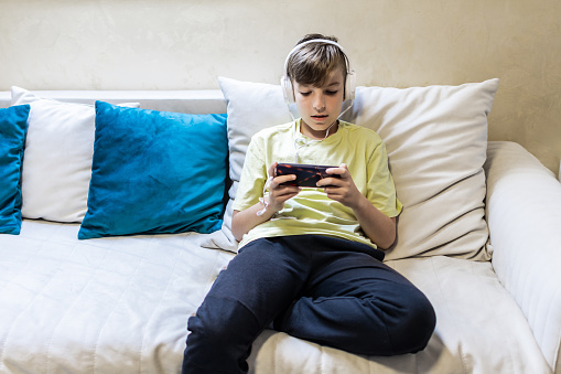 Schoolboy with headphones is playing video game on mobile phone at home.