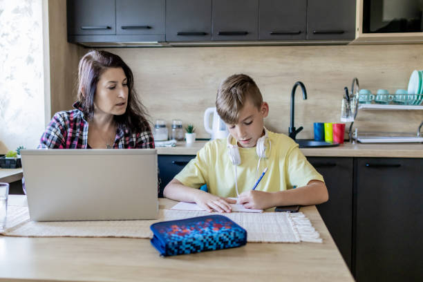 istock Homeschooling and freelancing. Mother is working on the laptop while her son is doing homework or studying. They are at home, sitting at kitchen table with school supplies and laptop. 1990364812