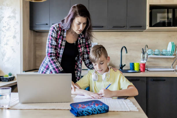 istock Homeschooling and freelancing. Mother is working on the laptop while her son is doing homework or studying. They are at home, sitting at kitchen table with school supplies and laptop. 1990366412