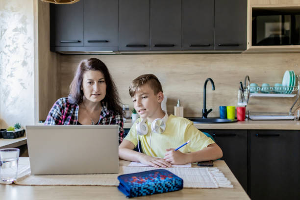 istock Homeschooling and freelancing. Mother is working on the laptop while her son is doing homework or studying. They are at home, sitting at kitchen table with school supplies and laptop. 1990372841