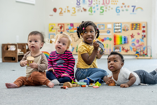 Four toddlers sit side-by-side on the floor of their daycare classroom as they pose for a portrait.  They are each dressed comfortably and have blocks out in front of them.