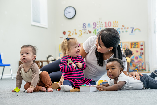A sweet little girl with Down Syndrome plays on the floor at Daycare with two of her friends and her female teacher.  She is dressed comfortably and appears happy as they play together with various toys.