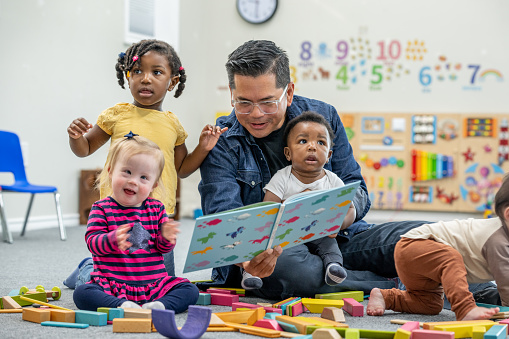 A male daycare teacher sits with a small group of toddlers on the floor of the classroom as he reads them a book.  They are each dressed comfortably and appear distracted by other toys around them.
