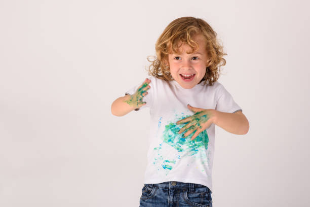 Delighted boy with curly hair smeared with colorful green paint looking away while standing on white background in light studio