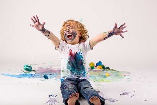 Full body of cheerful dirty boy sitting on messy painted floor with raised arms on white background in light studio