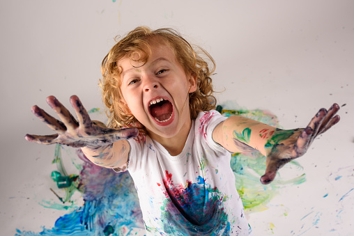 From above of funny boy covered with colorful paints looking at camera with excited face on white background in messy studio