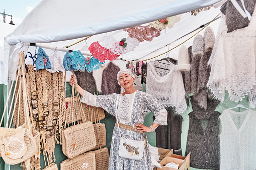 Irbit, Russia - August 11, 2023: Portrait of nice senior white woman, looking at camera, selling warm knitted items made of wool and fluff and crochet accessories at traditional fair.