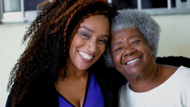 Happy portrait of adult African American daughter with arm around senior elderly gray-hair 80s mother. Close-up faces of joyful intergenerational women with arm around shoulder