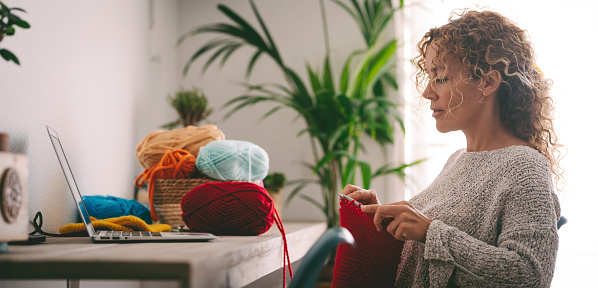 Woman sitting at desk crocheting a ball of red wool following a tutorial on her laptop learning the craft.Lifestyle and leisure concept