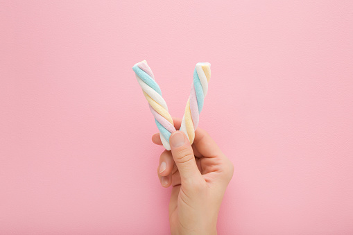 Young adult woman hand holding two colorful swirl soft marshmallow candies on light pink table background. Pastel color. Closeup. Sweet snack. Top down view.